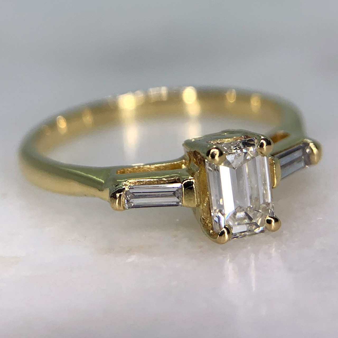 Stunning Baguette Diamond Trilogy Ring in 18ct Hallmarked yellow gold, London (1991). The ring is decorated with an 6 mm x 4.5 mm (approximate) emerald cut white diamond. The shoulders are embellished with two 3.5 mm x 1.3 mm baguette diamonds. All stones are claw set and weigh 0.60 carats of total diamond weight. This ring is in exquisite condition. 