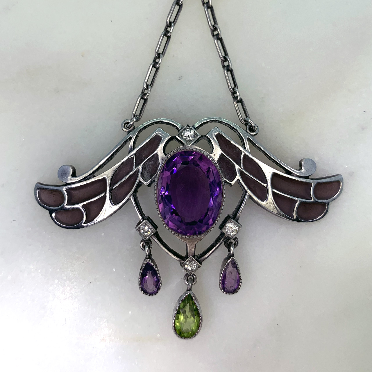 Exquisite Art Nouveau Silver Suffragette Pendant, set with Peridot, Amethysts and Diamonds. The central Amethyst measures approximately 13.3 mm x 10.8 mm, surrounded by four 3.8 mm diameter Diamonds, with three teardrop's hanging below (one Peridot, 2 Amethyst). All stones are rub-over set. The total pendant measures 50 mm x 70 mm (including the 33.50 mm chain and loop). On the back of the pendant is noted 'sterling'. 
